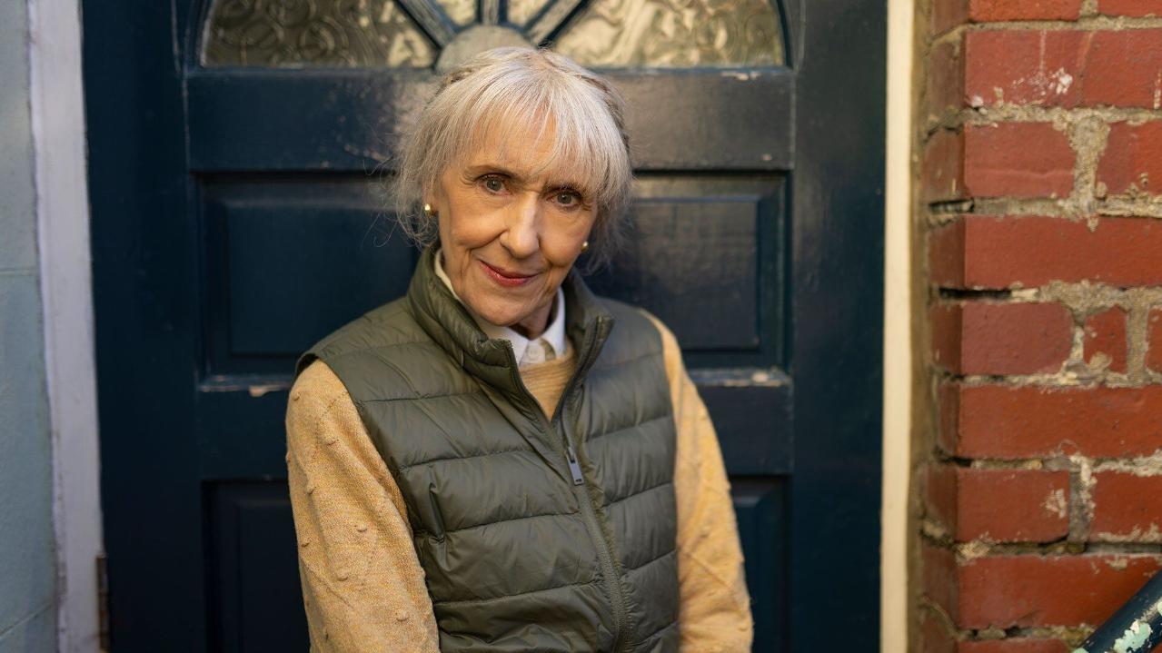 Anita Dobson as Mrs Flood smiles in front of a door in Doctor Who.