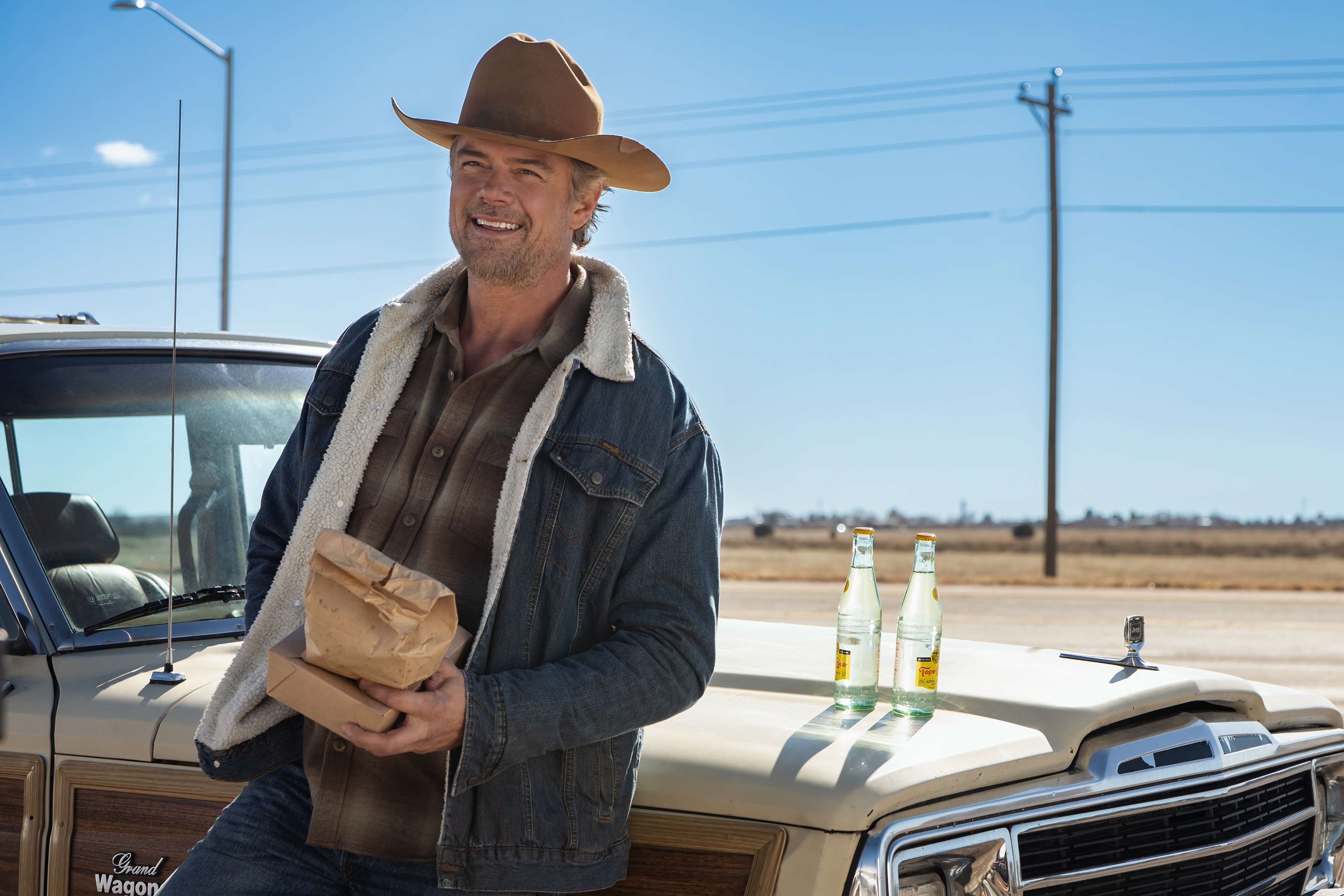 "Man in cowboy hat and denim jacket stands by vintage SUV holding paper bag on a sunny rural roadside. Two glass soda bottles sit on the car hood, with open field and power lines in the background."