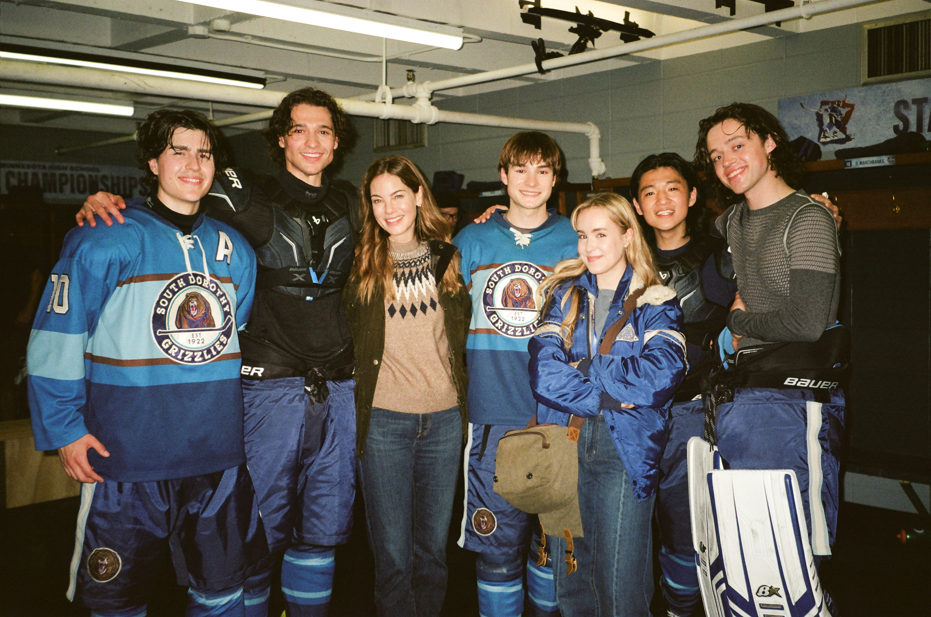 "Group of young hockey players in blue uniforms and two people in casual clothes smiling together inside a locker room, with benches and hockey equipment visible in the background."