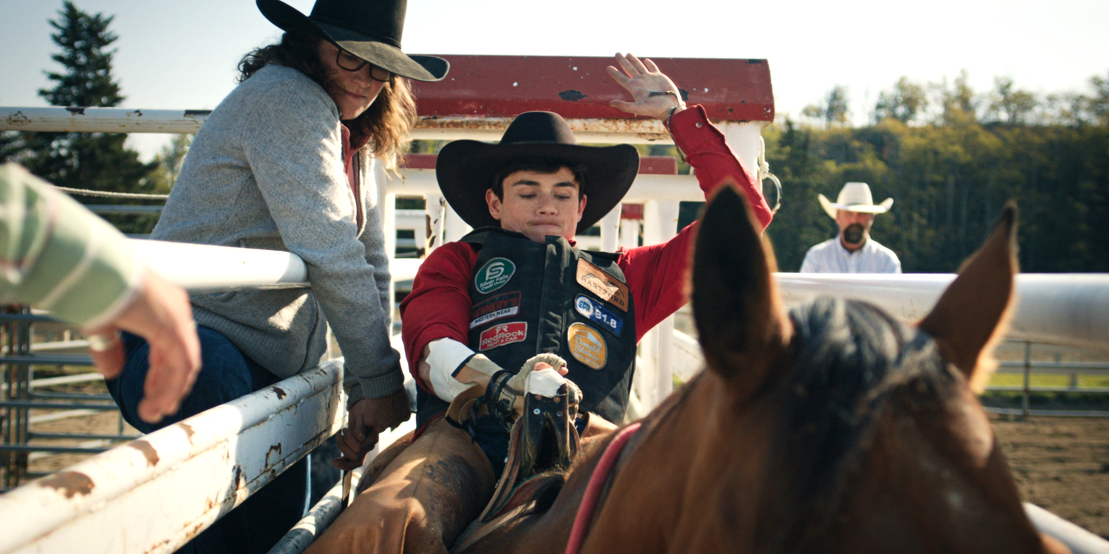 "Young rodeo rider in full gear prepares to ride a horse in an outdoor rodeo arena, with a helper nearby and spectators in western hats in the background on a sunny day."